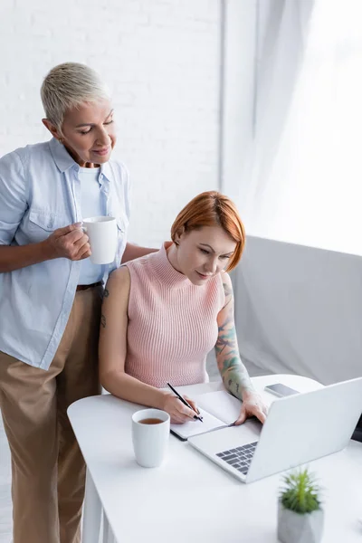 Lesbian woman holding cup of tea near girlfriend writing in notebook while working at home — Stock Photo