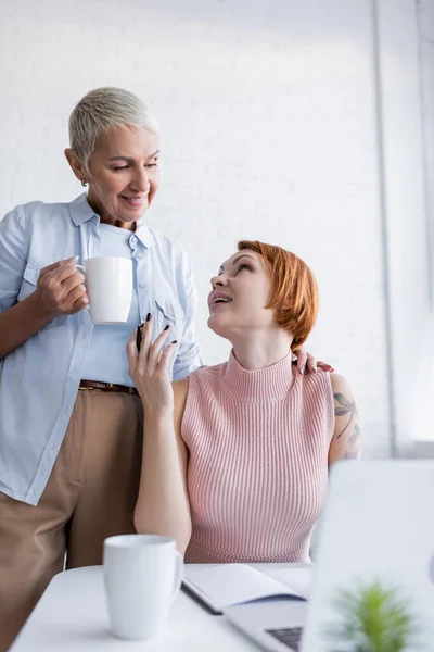 Mujer lesbiana hablando con una novia sonriente mientras está sentada cerca de una computadora portátil borrosa en casa - foto de stock