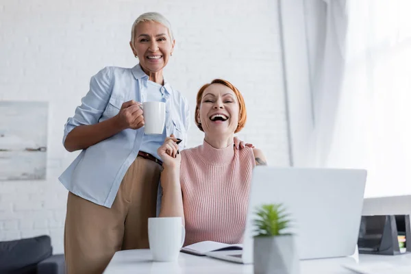 Eccitate donne lesbiche ridendo della fotocamera vicino al computer portatile offuscato sul tavolo — Foto stock