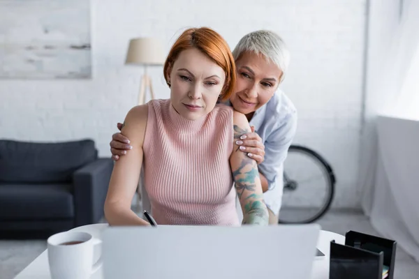 Mujer lesbiana positiva sonriendo cerca de novia pensando cerca borrosa portátil - foto de stock