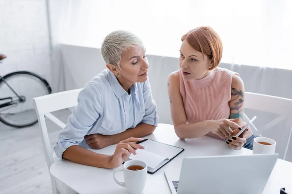 Tätowierte Frau zeigt auf Laptop, während sie mit Freundin in der Nähe von Laptop und Notebook sitzt — Stockfoto