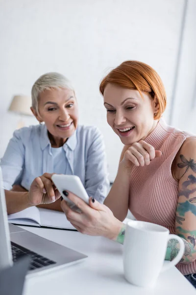 Souriant lesbienne femme pointant vers smartphone dans la main de petite amie tatouée assis près d'un ordinateur portable et tasse de thé — Photo de stock