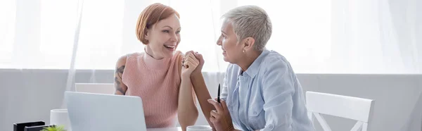 Alegres mujeres lesbianas mirándose mientras se toman de la mano cerca de la computadora portátil, pancarta - foto de stock