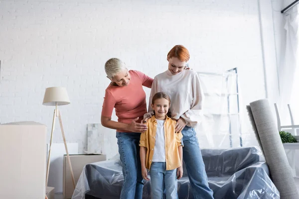 Happy preteen girl smiling at camera near same sex parents in new apartment — Stock Photo