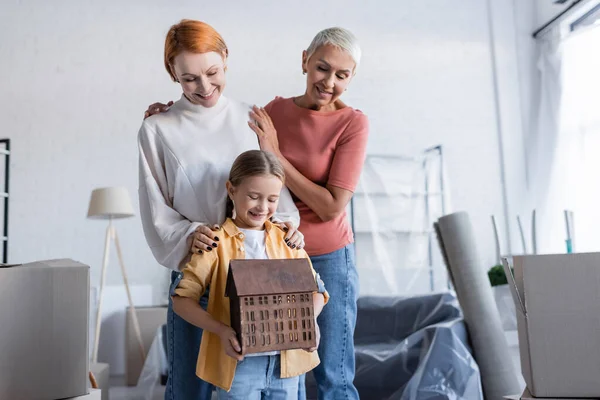 Cheerful girl holding house model near same sex mothers in new home — Stock Photo