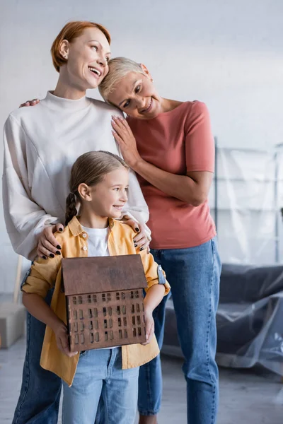 Preteen girl holding house model near pleased same sex parents in new home — Stock Photo