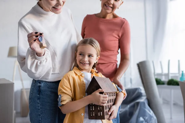 Cheerful girl holding house model near same sex parents with key from new apartment — Stock Photo