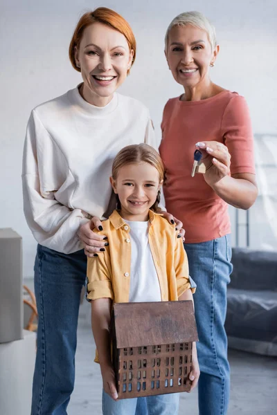 Lesbian couple looking at camera near happy adopted daughter holding house model — Stock Photo