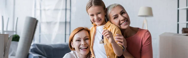 Mulheres lésbicas alegres olhando para a câmera perto da filha adotada segurando a chave do novo apartamento, banner — Fotografia de Stock