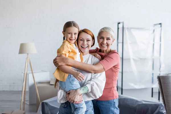 Joyful lesbian couple with adopted daughter smiling at camera in new apartment — Stock Photo