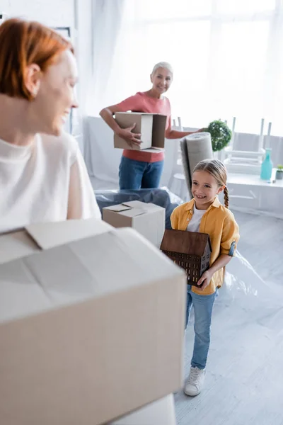 Joyful girl carrying house model near blurred same sex parents with carton boxes — Stock Photo