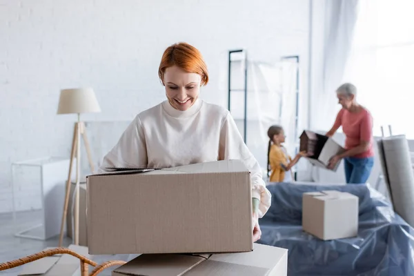 Femme lesbienne souriante tenant paquet de carton près de la famille floue dans un nouvel appartement — Photo de stock