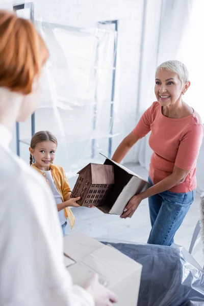 Smiling woman holding carton package near adopted daughter with house model and blurred girlfriend — Stock Photo