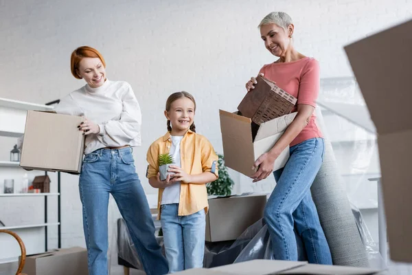 Lesbian women holding cardboard boxes near happy child with flowerpot in new apartment — Stock Photo
