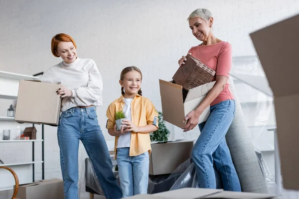 Lesbiennes femmes tenant des boîtes en carton près de heureux enfant avec pot de fleurs dans le nouvel appartement — Photo de stock