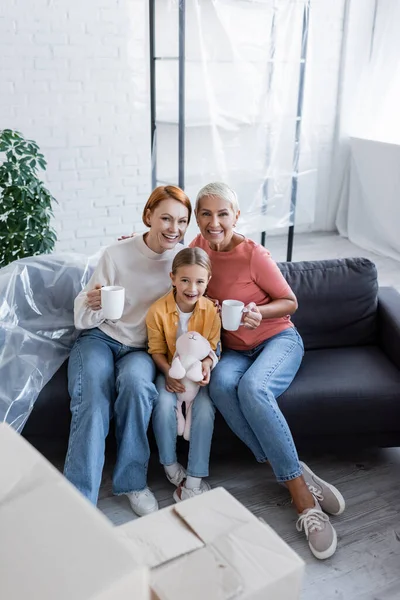 Feliz lesbiana mujer con tazas de té y adoptivo chica con juguete conejito sentado en sofá en nuevo hogar - foto de stock