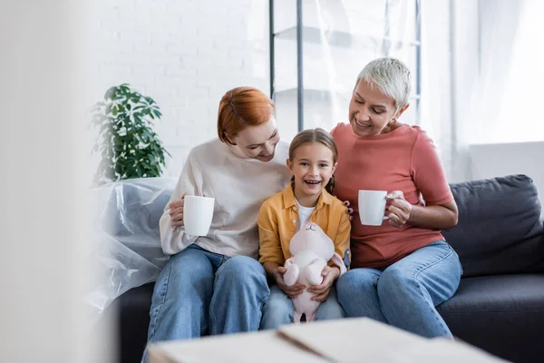 Alegre chica con juguete conejito mirando la cámara cerca del mismo sexo madres sentadas con tazas de té en nuevo hogar - foto de stock