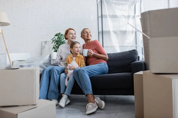 Веселые лесбиянки с приемной дочерью, смотрящие на диван в новой квартире — стоковое фото