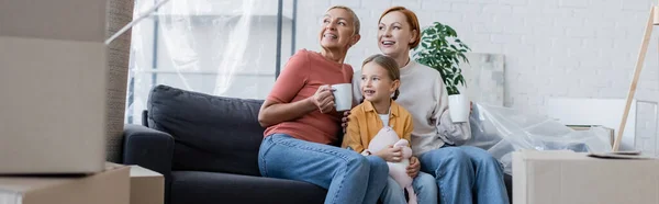 Femmes lesbiennes heureuses et fille adoptive assise sur le canapé dans un nouvel appartement et regardant ailleurs, bannière — Photo de stock