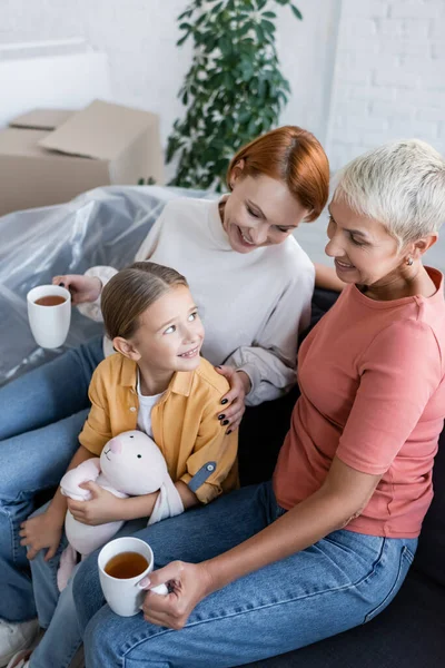 Mulheres lésbicas com xícaras de chá e menina com coelho de brinquedo sorrindo no sofá no novo apartamento — Fotografia de Stock