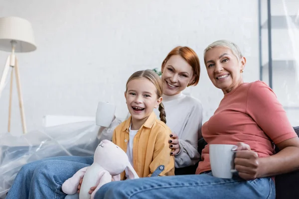 Cheerful lesbian women and adopted girl with toy bunny looking at camera in new home — Stock Photo