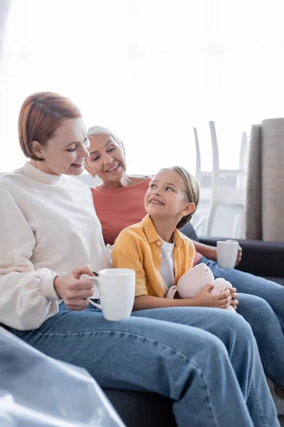 Feliz chica con juguete conejito sentado cerca del mismo sexo padres en nuevo hogar - foto de stock