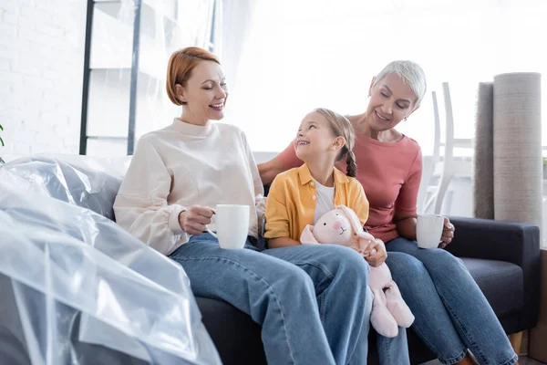 Fröhliches Mädchen sitzt auf Couch neben gleichgeschlechtlichen Eltern in neuer Wohnung — Stockfoto