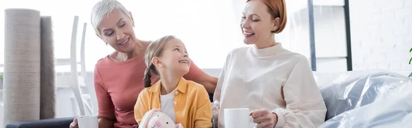 Joyful lesbian women with adopted girl smiling at each other in new home, banner — Stock Photo