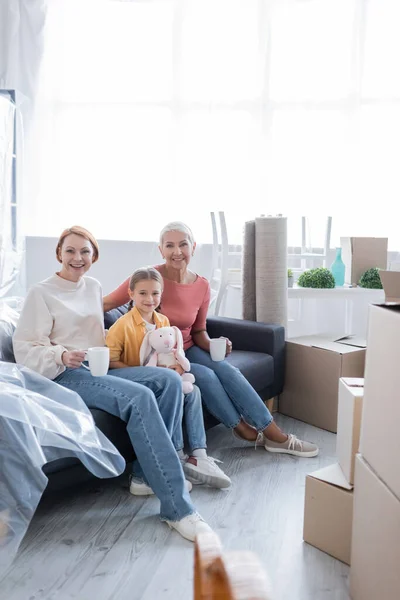 Preteen girl with soft toy and lesbian women with tea cups smiling on sofa in new home — Stock Photo