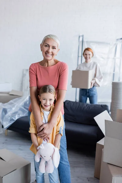 Glückliche lesbische Frau mit Adoptivtochter lächelt in der Nähe von Kartons im neuen Zuhause in die Kamera — Stockfoto