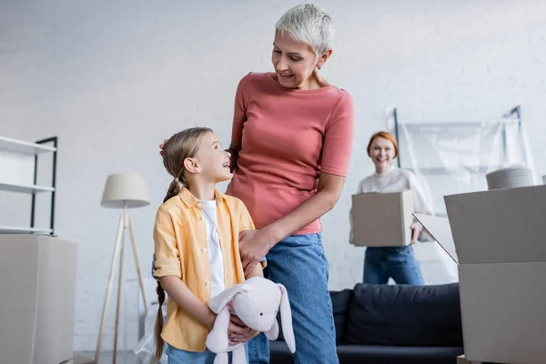 Lesbiana mujer y adoptiva chica con juguete conejito sonriendo el uno al otro en nuevo hogar - foto de stock