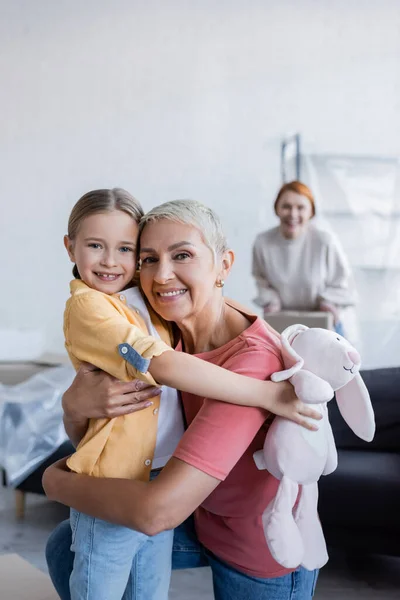 Cheerful lesbian woman embracing adopted daughter and looking at camera in new home — Stock Photo