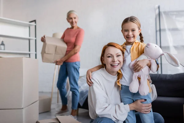 Glückliche lesbische Frau umarmt Adoptivtochter mit Spielzeughasen in neuer Wohnung — Stockfoto