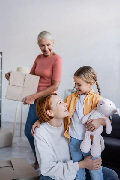 Happy lesbian woman looking at adopted girl holding toy bunny in new apartment — Stock Photo