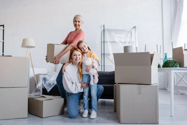 Joyful same sex couple with adopted daughter looking at camera near carton packages in new home — Stock Photo