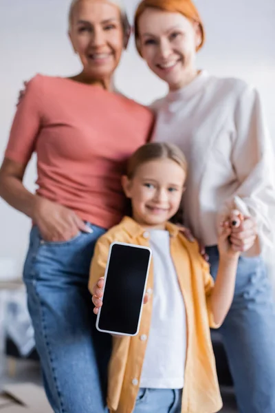 Foco seletivo do smartphone com tela em branco na mão da menina sorrindo perto dos pais do mesmo sexo — Fotografia de Stock