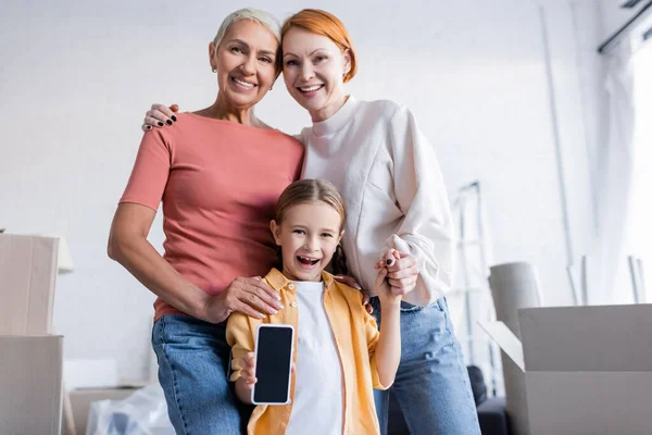 Positives lesbisches Paar umarmt Kind zu Hause mit Smartphone und Kartonschachteln — Stockfoto