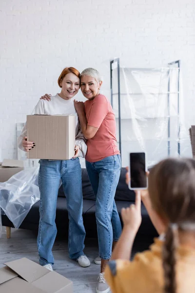 Lächelndes lesbisches Paar hält Karton in der Hand, während Tochter zu Hause ein Handy-Foto macht — Stockfoto