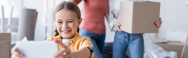 Enfant souriant prenant selfie près des mères floues avec boîte en carton à la maison, bannière — Photo de stock