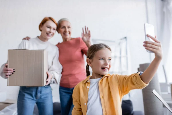 Smiling kid having video call near blurred mothers with carton box at home — Stock Photo