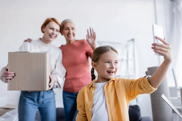 Lächelndes Kind bei Videoanruf in der Nähe verschwommener Mütter mit Karton zu Hause — Stockfoto