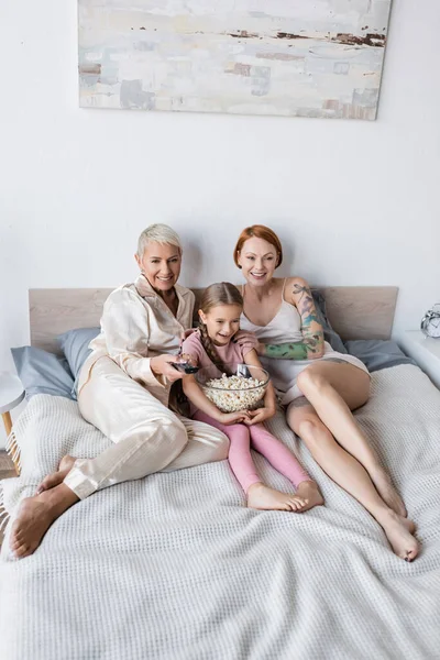 Mulheres lésbicas positivas assistindo tv perto de criança com pipoca em casa — Fotografia de Stock