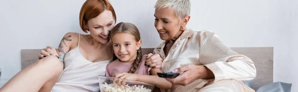Ragazza sorridente che tiene i popcorn vicino alle mamme lesbiche con telecomando sul letto, banner — Foto stock