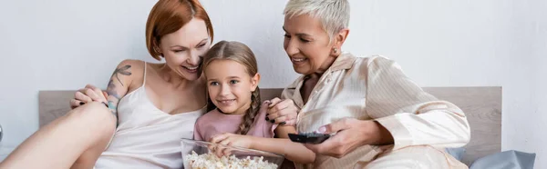 Sorrindo menina segurando pipocas perto de mães lésbicas com controle remoto na cama, banner — Fotografia de Stock