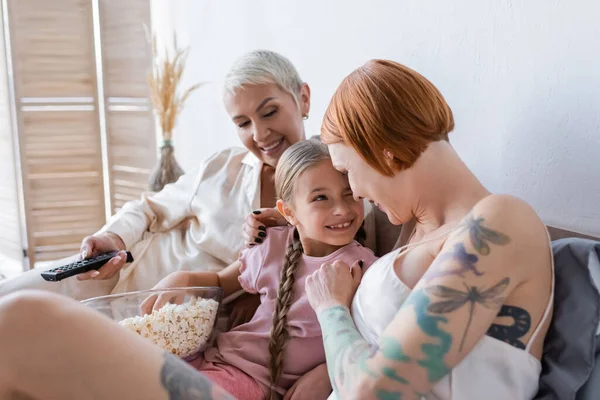 Woman hugging child with popcorn near girlfriend with remote controller at home — Stock Photo