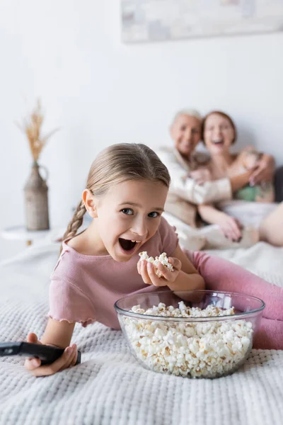 Excited girl holding popcorn and remote controller near blurred mothers on bed — Stock Photo