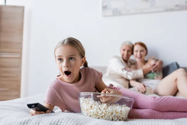 Impresionado niño viendo la televisión cerca de palomitas de maíz y las madres borrosas en la cama - foto de stock