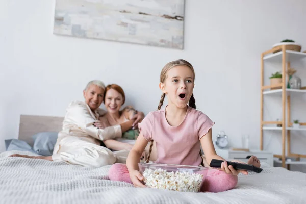 Excited girl holding remote controller and popcorn near blurred mothers at home — Stock Photo
