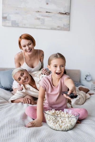 Positive girl clicking channels near popcorn and blurred mothers on bed — Stock Photo