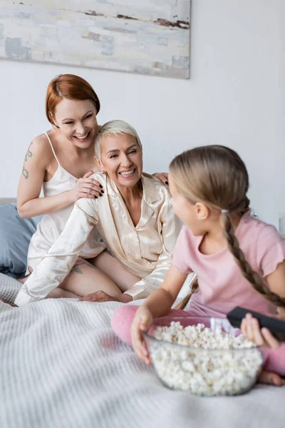 Feliz pareja lesbiana abrazándose cerca del niño con mando a distancia y palomitas de maíz en la cama - foto de stock